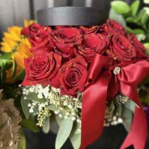 A luxury black hatbox filled with red roses, gold sparkle, greenery, and a stunning satin bow. Perfect for birthdays, romantic gifts, anniversaries, and celebrations.