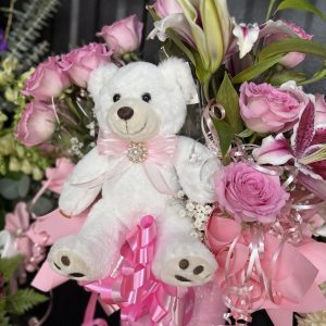 Pink rose and lily arrangement with a plush teddy bear, jeweled bow, and cascading ribbons. Perfect for birthdays, quinceañeras, baby showers, and special celebrations.
