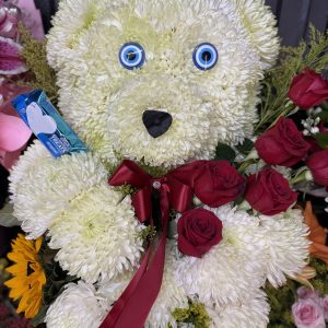 A handcrafted floral teddy bear made with white mums, red roses, and a gold crown. A unique and adorable arrangement perfect for birthdays, quinceañeras, and special celebrations.